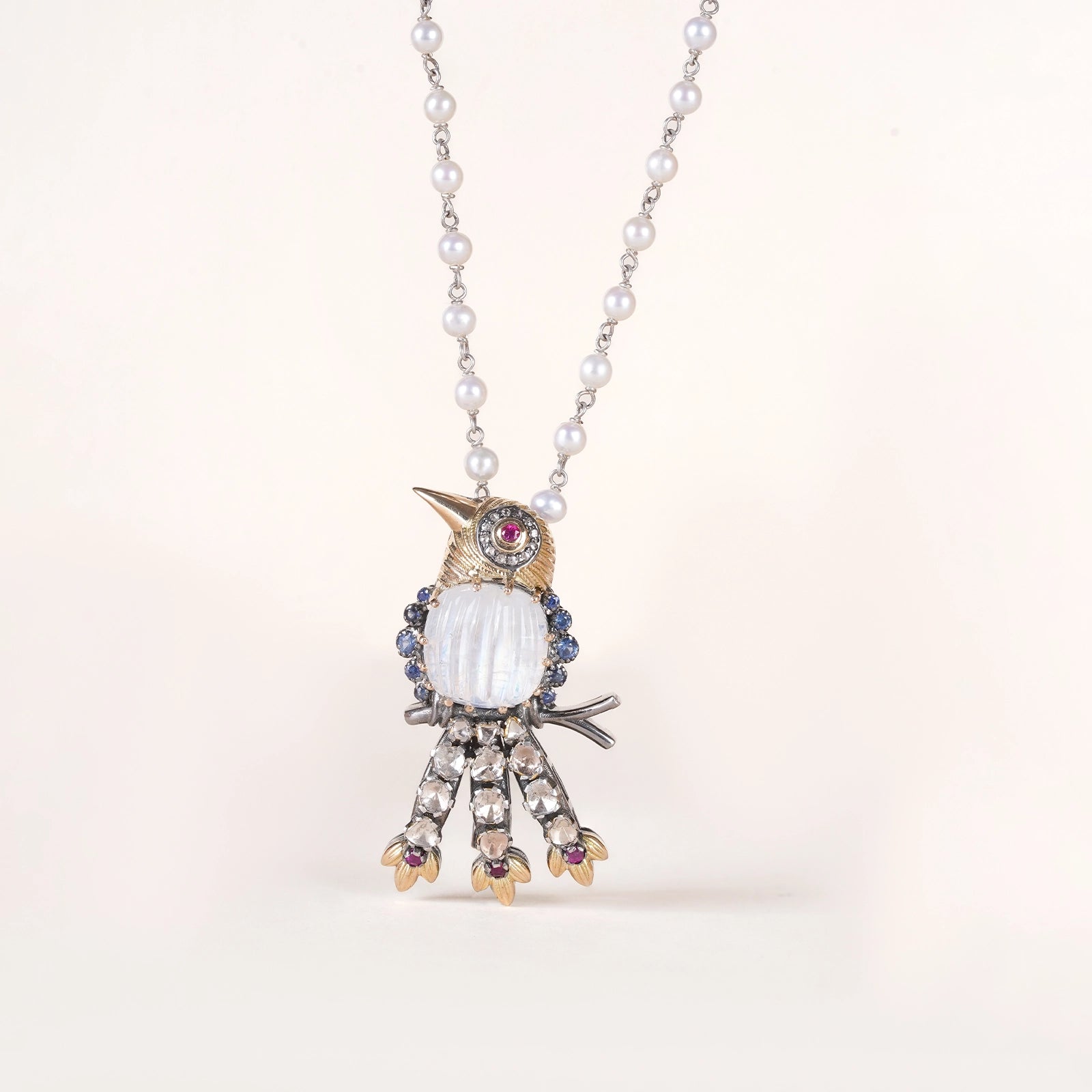 Lovebird Sky Brooch featuring a carved moonstone bird with blue sapphire detailing, ruby eye, uncut diamond tail feathers, and pearl accented chain in gold.